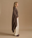 A woman stands in profile wearing a brown open-front knit cardigan draped over cream wide-leg trousers and dark slip-on shoes, against a plain warm beige studio background.