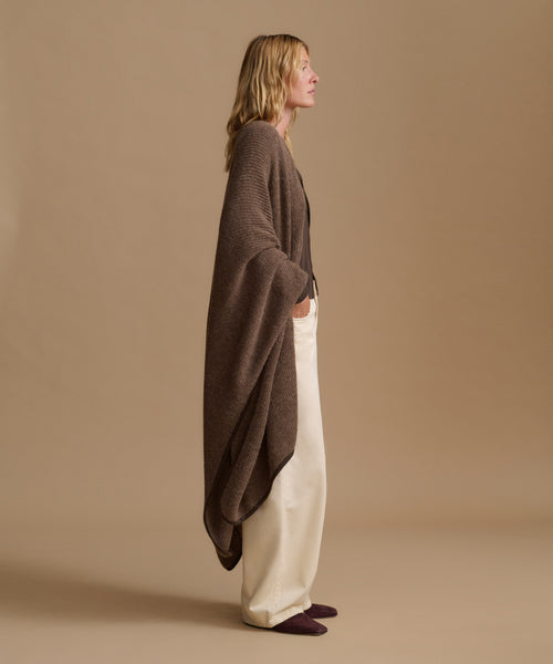 A woman stands in profile wearing a brown open-front knit cardigan draped over cream wide-leg trousers and dark slip-on shoes, against a plain warm beige studio background.