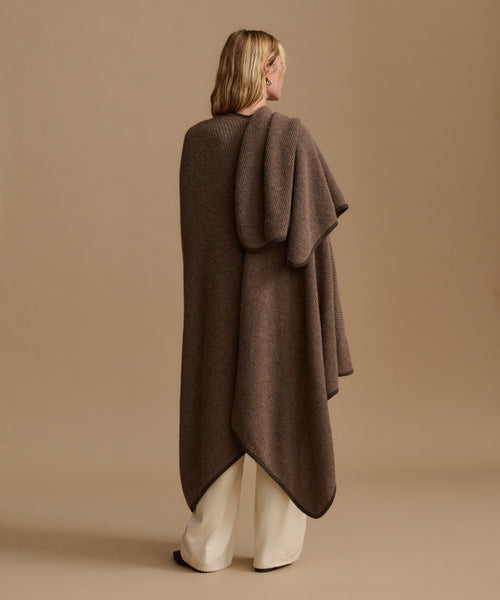 A woman shown from the back wearing a long, draped brown knit poncho or cape over cream wide-leg pants, standing against a plain beige studio background.