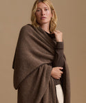 A woman modeling a chunky knit brown shawl wrap draped over her shoulders, paired with a dark brown long-sleeve top and cream pants, against a solid beige studio background.
