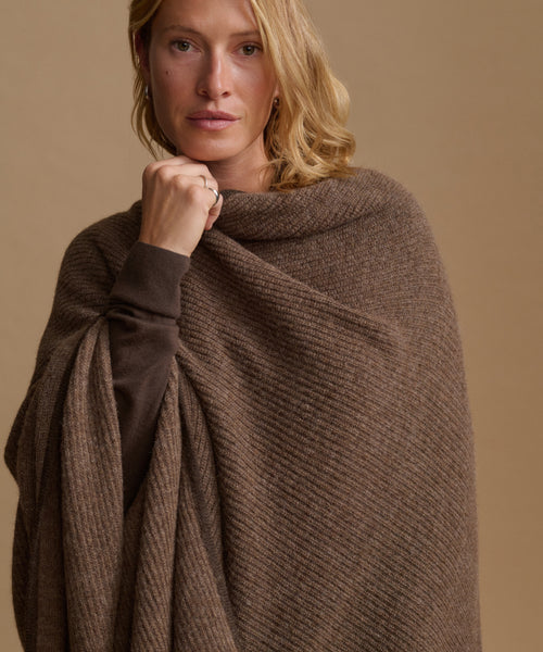 A woman wearing a brown ribbed knit wrap or poncho over a dark brown top, photographed against a plain warm beige studio background.