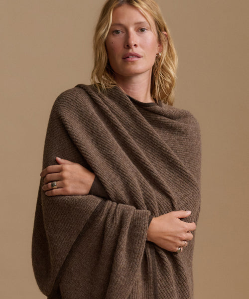 A woman wearing a brown textured knit wrap or poncho draped over her shoulders, posed against a solid warm beige background.