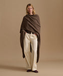 A woman stands against a warm beige studio background wearing a dark brown draped wrap or poncho over cream wide-leg trousers and dark slip-on shoes.