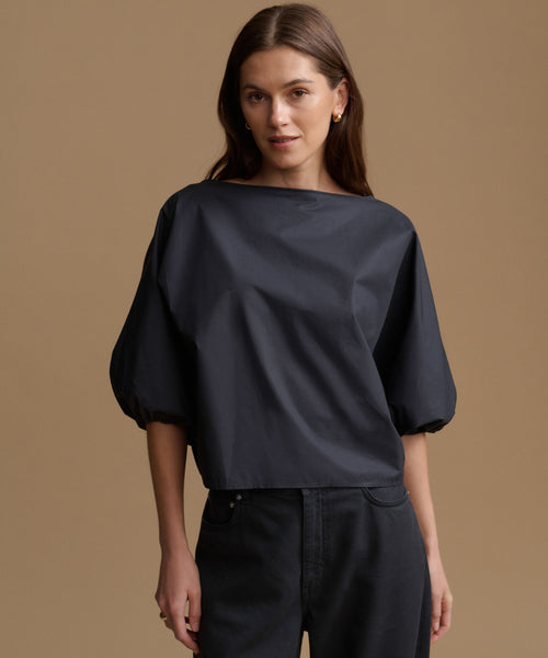 A woman modeling a black off-the-shoulder blouse with voluminous puff sleeves and dark charcoal pants, set against a solid warm brown studio background.