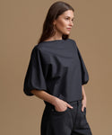 A woman wearing a black off-the-shoulder puff-sleeve top and dark jeans, standing against a solid warm brown studio background.