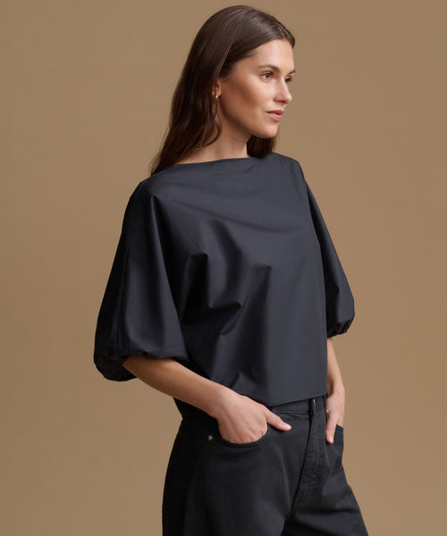 A woman wearing a black off-the-shoulder puff-sleeve top and dark jeans, standing against a solid warm brown studio background.