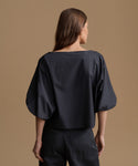 A woman shown from the back wearing a loose, wide-neck black top with voluminous elbow-length sleeves and black pants, standing against a plain warm tan studio background.