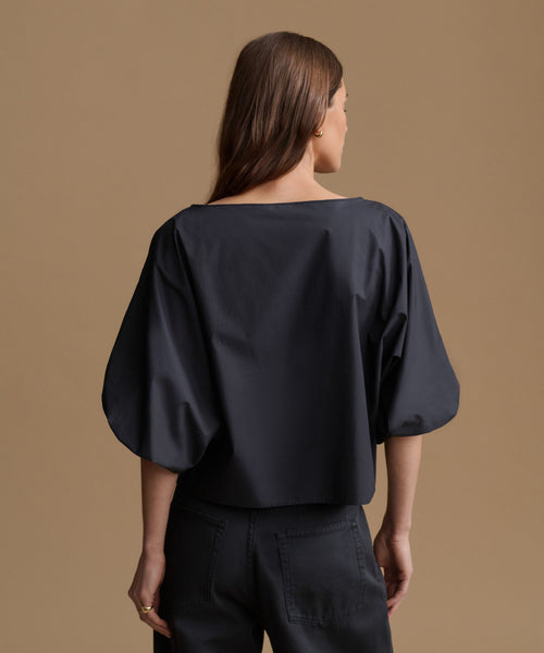 A woman shown from the back wearing a loose, wide-neck black top with voluminous elbow-length sleeves and black pants, standing against a plain warm tan studio background.
