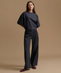 A woman modeling a black two-piece outfit consisting of a wide-neck, short-sleeved top and matching wide-leg trousers, paired with dark burgundy flats, against a solid beige studio background.
