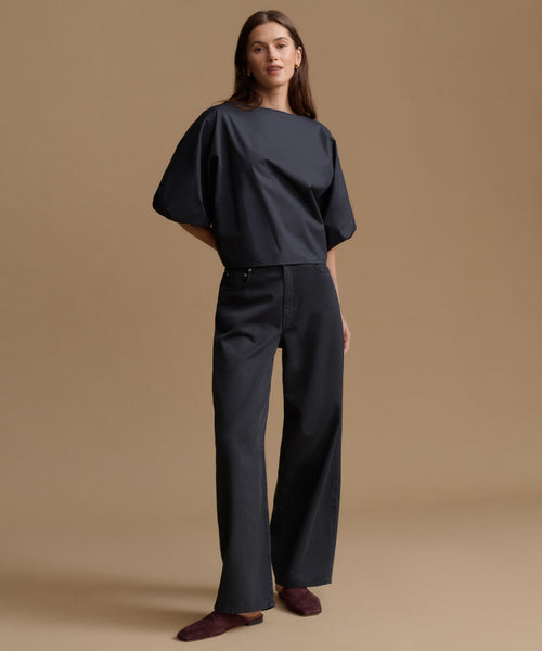 A woman modeling a black two-piece outfit consisting of a wide-neck, short-sleeved top and matching wide-leg trousers, paired with dark burgundy flats, against a solid beige studio background.