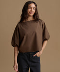 A woman wearing a brown short-sleeve top with puff sleeves and black pants, standing against a solid tan background.