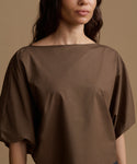 A close-up of a woman wearing a loose-fitting, wide-neck top in a solid medium brown color, set against a warm beige studio background.