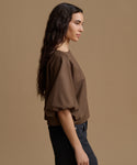 A woman in side profile wearing a brown puff-sleeve blouse and dark jeans, set against a solid warm brown studio background.