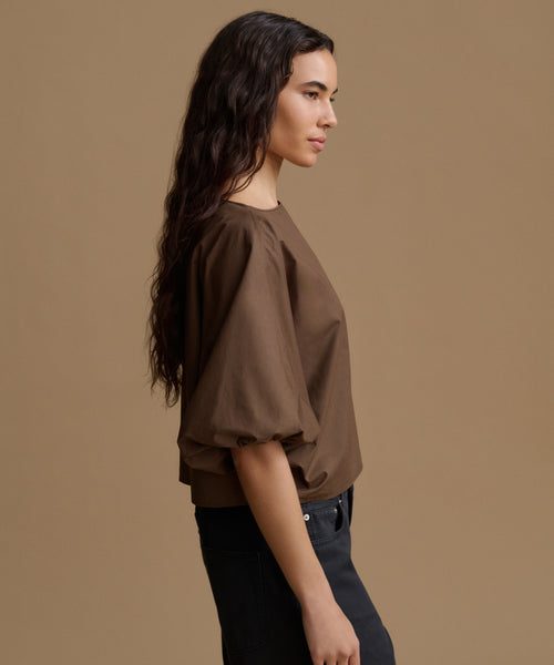 A woman in side profile wearing a brown puff-sleeve blouse and dark jeans, set against a solid warm brown studio background.