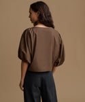 A woman shown from the back wearing a brown cropped blouse with voluminous puff sleeves and dark wide-leg trousers, set against a solid warm beige studio background.