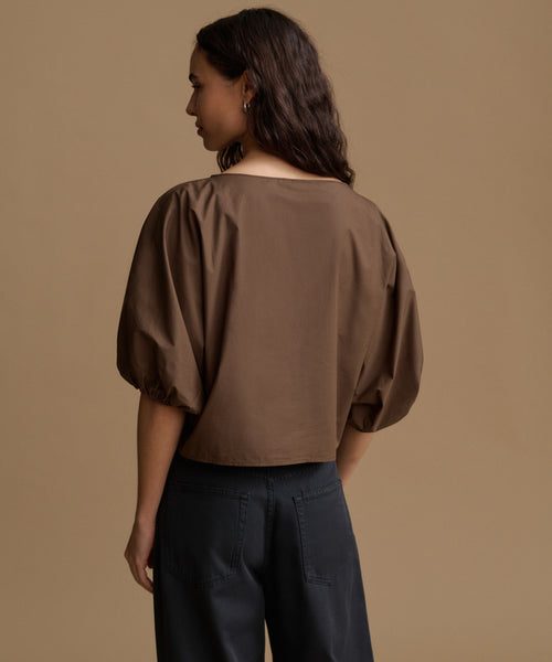 A woman shown from the back wearing a brown cropped blouse with voluminous puff sleeves and dark wide-leg trousers, set against a solid warm beige studio background.