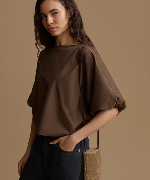A woman wearing a brown short-sleeve top with voluminous elbow-length sleeves and dark pants, standing against a solid tan background.