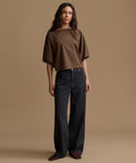 A woman stands against a plain warm beige studio background wearing a loose brown short-sleeve top and dark charcoal wide-leg pants.