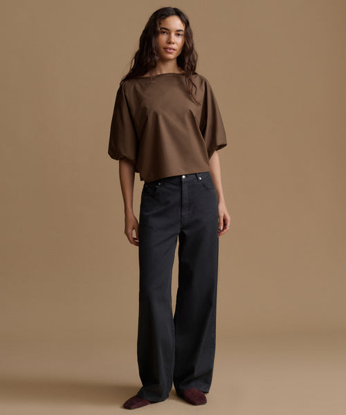 A woman stands against a plain warm beige studio background wearing a loose brown short-sleeve top and dark charcoal wide-leg pants.