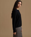 A woman wearing a black long-sleeve knit sweater and striped trousers, shown in profile against a solid tan studio background.