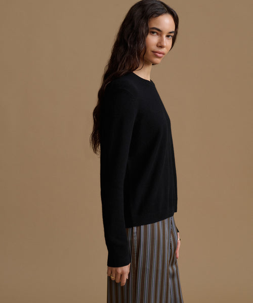 A woman wearing a black long-sleeve knit sweater and striped trousers, shown in profile against a solid tan studio background.