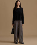 A woman stands against a warm beige studio background wearing a black long-sleeve top and wide-leg striped trousers in brown, gray, and beige tones. She holds a small black handbag.