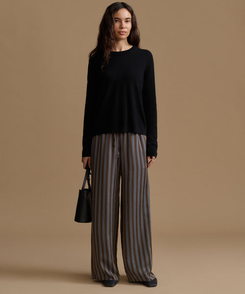 A woman stands against a warm beige studio background wearing a black long-sleeve top and wide-leg striped trousers in brown, gray, and beige tones. She holds a small black handbag.