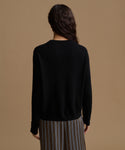 A woman shown from the back wearing a black long-sleeve knit sweater and striped trousers against a solid tan background.