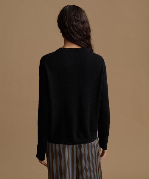 A woman shown from the back wearing a black long-sleeve knit sweater and striped trousers against a solid tan background.
