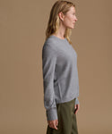 A woman in side profile wearing a light gray long-sleeve knit sweater and olive green trousers, set against a solid warm tan studio background.