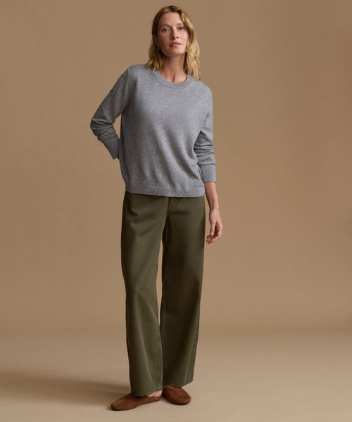 A woman stands against a warm beige studio background wearing a light gray long-sleeve knit sweater, olive green wide-leg trousers, and brown flat shoes.