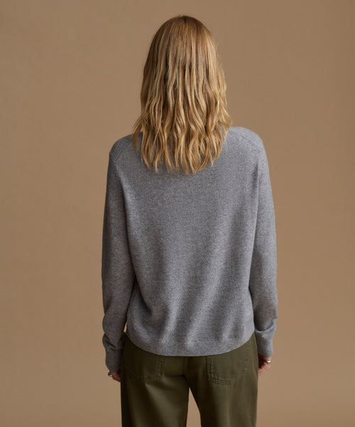 A woman viewed from behind wearing a heather gray long-sleeve knit sweater and olive green pants, standing against a solid warm tan studio background.