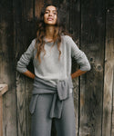 A woman wearing a light gray knit long-sleeve top and matching gray wide-leg pants, standing with hands on hips against a dark rustic wooden plank wall.