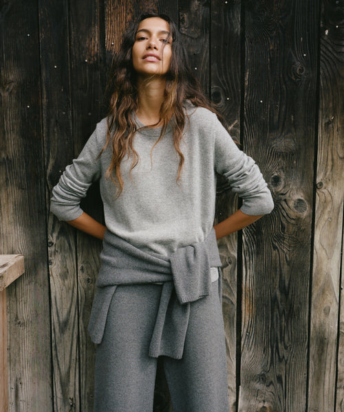 A woman wearing a light gray knit long-sleeve top and matching gray wide-leg pants, standing with hands on hips against a dark rustic wooden plank wall.