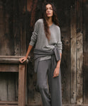 A woman wearing a heather gray knit sweater and matching wide-leg knit pants, standing against a rustic dark wood plank wall and leaning on a wooden bench.