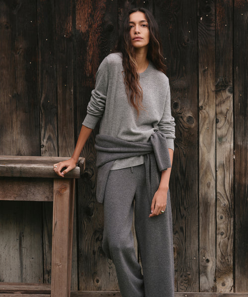 A woman wearing a heather gray knit sweater and matching wide-leg knit pants, standing against a rustic dark wood plank wall and leaning on a wooden bench.