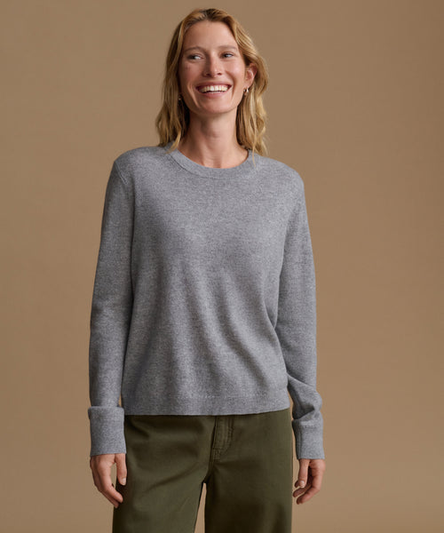 A woman smiling and wearing a light gray long-sleeve crewneck sweater with olive green pants, standing against a solid tan studio background.