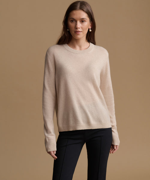 A woman modeling a light beige crewneck knit sweater with long sleeves, paired with black pants, against a solid warm beige studio background.