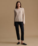 A woman stands against a warm beige studio background wearing a light cream crewneck sweater, black cropped trousers, and dark slip-on shoes.