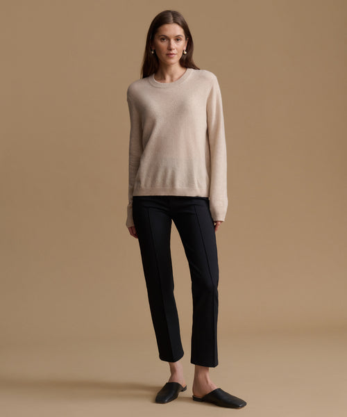 A woman stands against a warm beige studio background wearing a light cream crewneck sweater, black cropped trousers, and dark slip-on shoes.