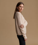 A woman modeling a light beige knit sweater with long sleeves, shown from a side-back angle against a solid tan studio background.