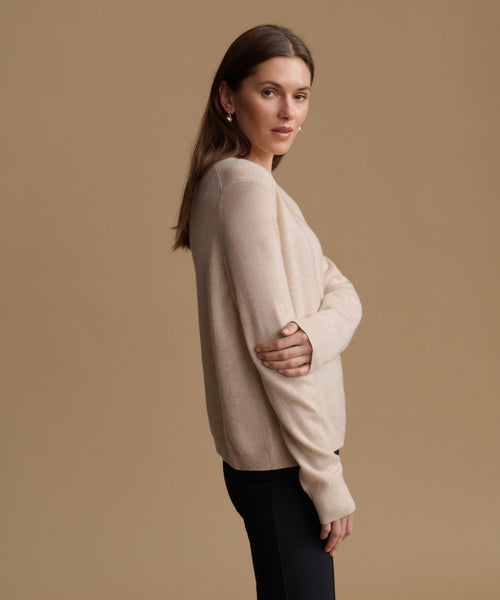 A woman modeling a light beige knit sweater with long sleeves, shown from a side-back angle against a solid tan studio background.
