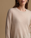 A close-up of a woman wearing a soft, light beige crewneck sweater against a solid warm beige background.