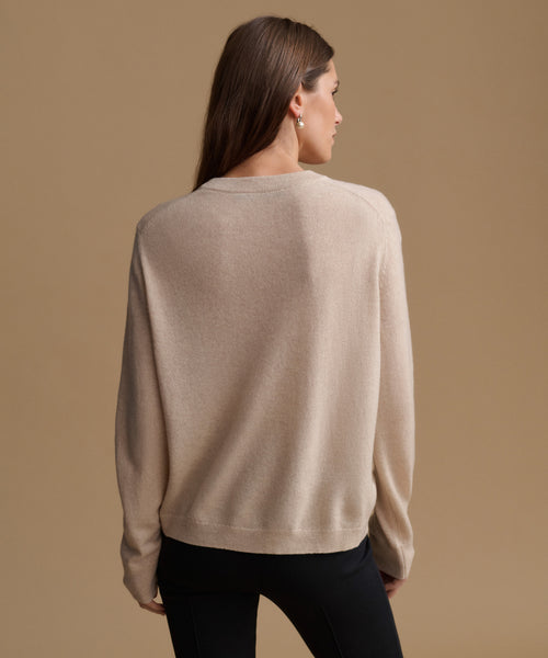 A woman shown from the back wearing a light beige knit sweater and black pants against a solid tan studio background.
