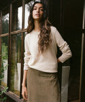 A woman with long wavy brown hair stands by a dark-framed window, wearing a cream knit crewneck sweater and a taupe skirt.