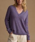 A woman wearing a heathered purple V-neck knit sweater and cream pants, standing against a solid tan background.