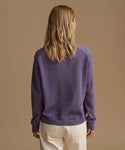 A woman viewed from behind wearing a heathered purple knit sweater and cream trousers, standing against a solid tan studio background.