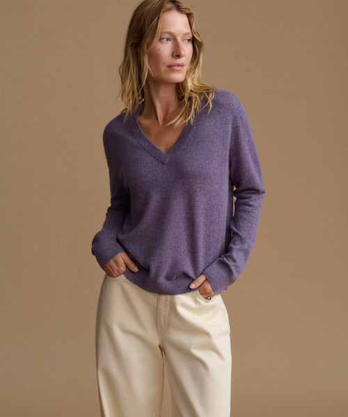 A woman wearing a heathered purple long-sleeve V-neck sweater and cream-colored pants, standing against a solid tan studio background.