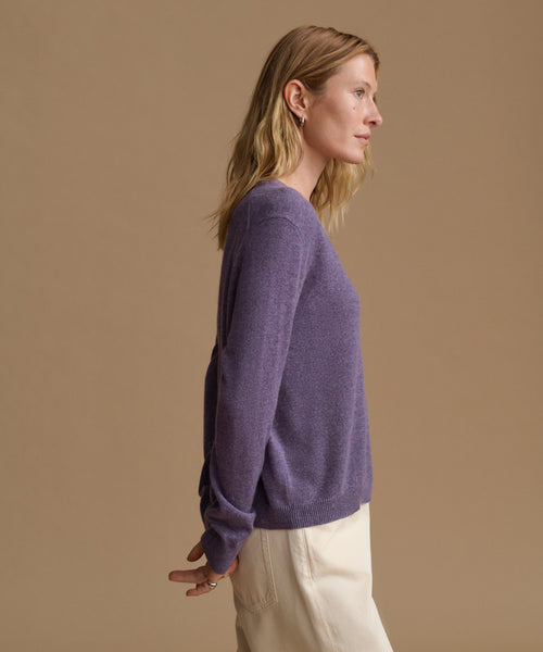 A woman in side profile wearing a heathered purple knit sweater and cream trousers, set against a solid warm beige studio background.