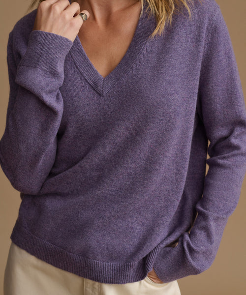 A close-up of a person wearing a heathered purple V-neck knit sweater with long sleeves and ribbed trim, paired with light beige pants against a warm beige background.